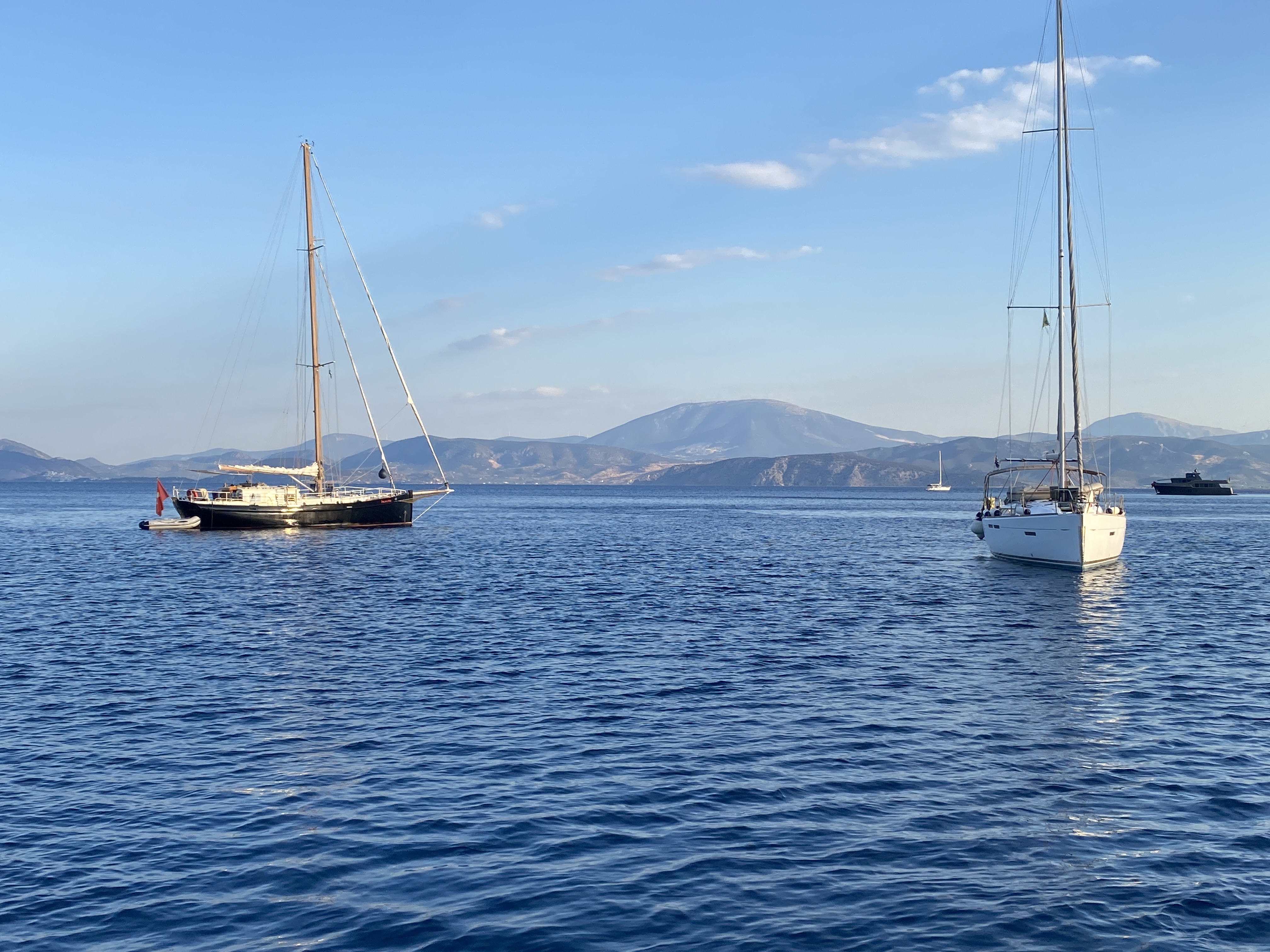 Two sailing yachts at anchor on blue water beneath a mountainous coastline